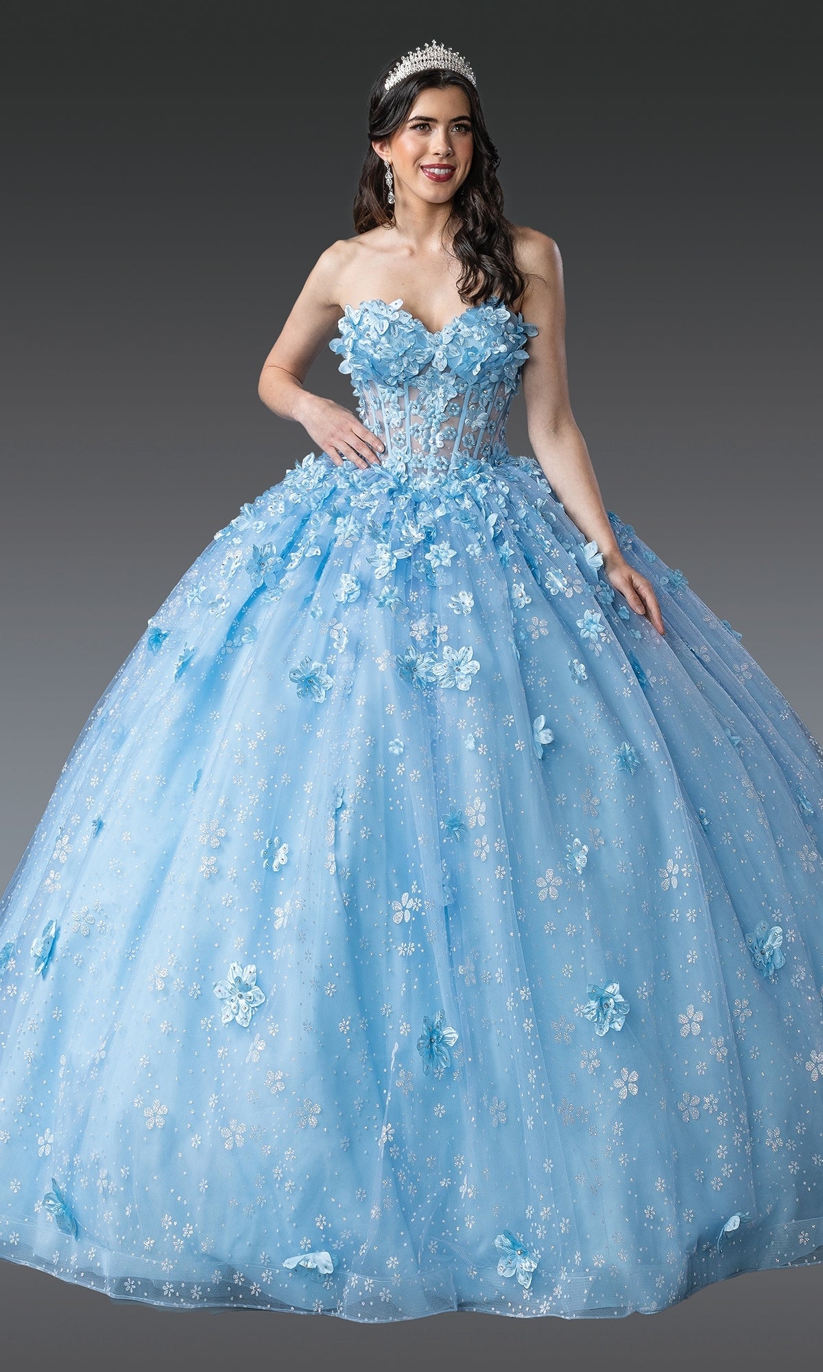 A woman in the Blue Quinceanera Dress 1972 by Dancing Queen, featuring 3D floral appliquรฉs and a tiara, stands against a plain gray background.